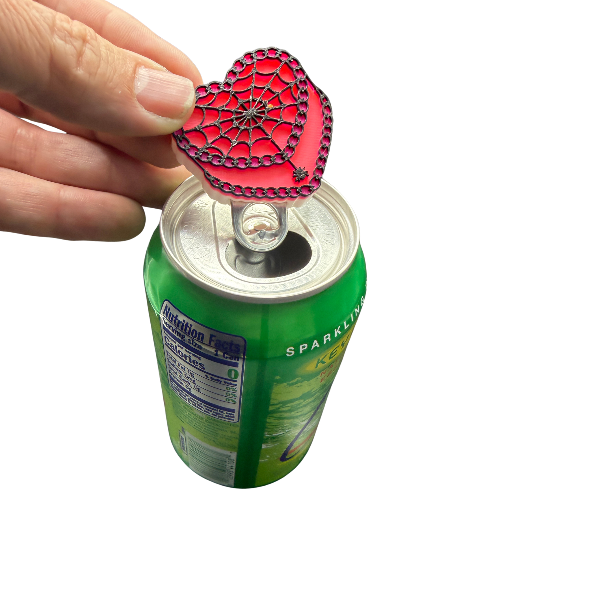 A hand holding a red heart-shaped silicone can opener with a spiderweb design, shown opening a green beverage can against a white background.